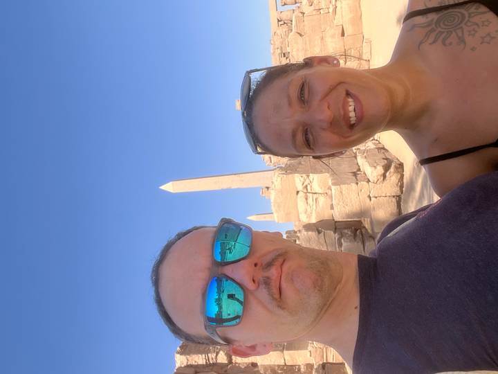 Couple stands before ancient sandstone obelisks under a cloudless blue sky.
