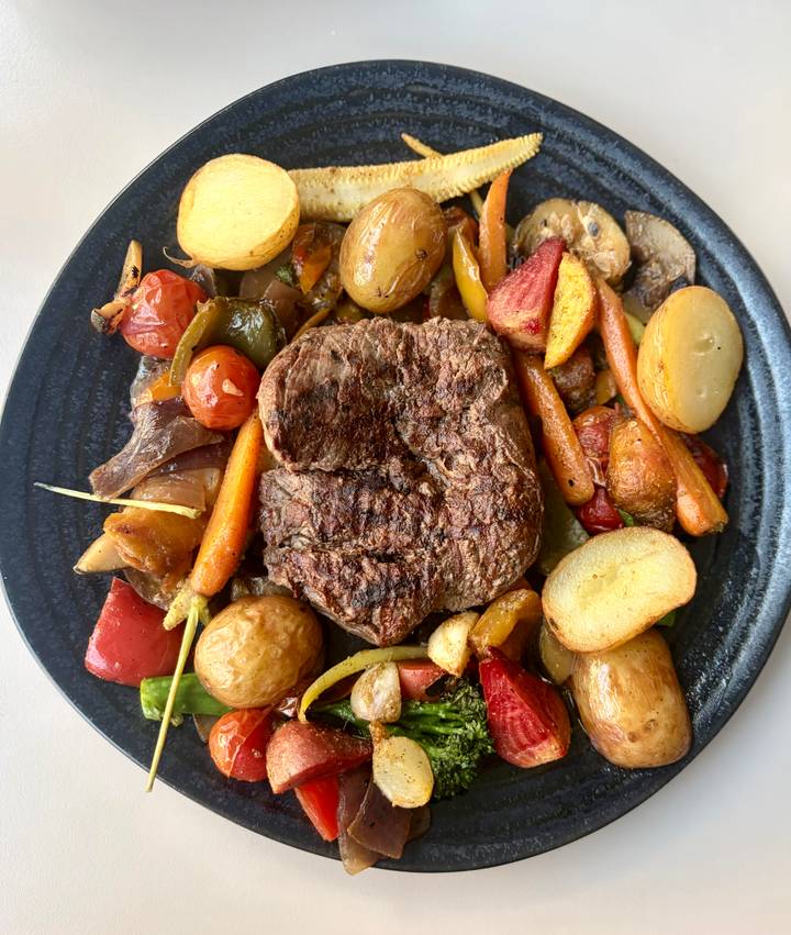 Grilled beef steak surrounded by roasted vegetables and baby potatoes on a dark plate.