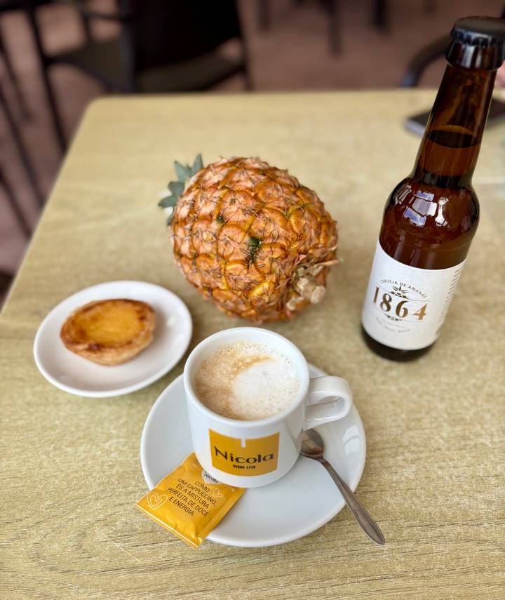Cappuccino, whole pineapple, pastel de nata and craft beer arranged on a table.