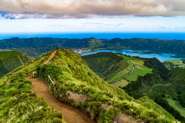 Scenic hiking trail follows a lush green ridge overlooking crater lake and distant ocean.