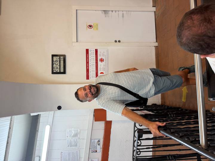 A smiling man leans casually against a railing inside a station area with a wall clock and signage behind him.