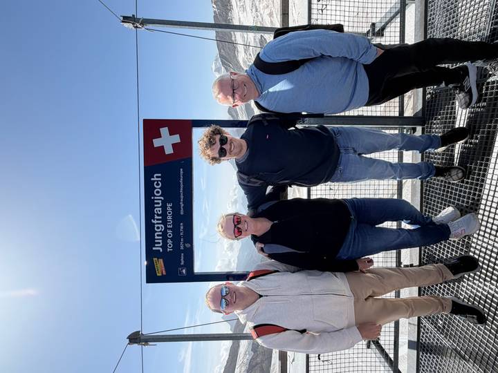 Four travellers pose in front of a sign reading 'Jungfraujoch Top of Europe' with snowy Alpine peaks in the background.