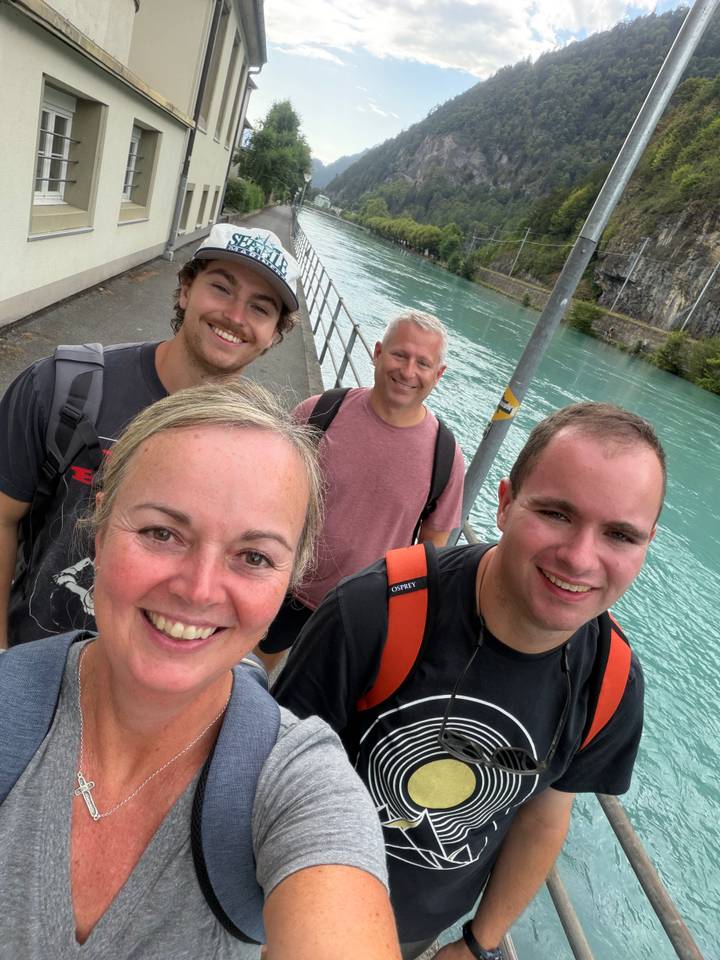 A close-up selfie of four smiling travellers walking beside a bright turquoise river with rocky banks.