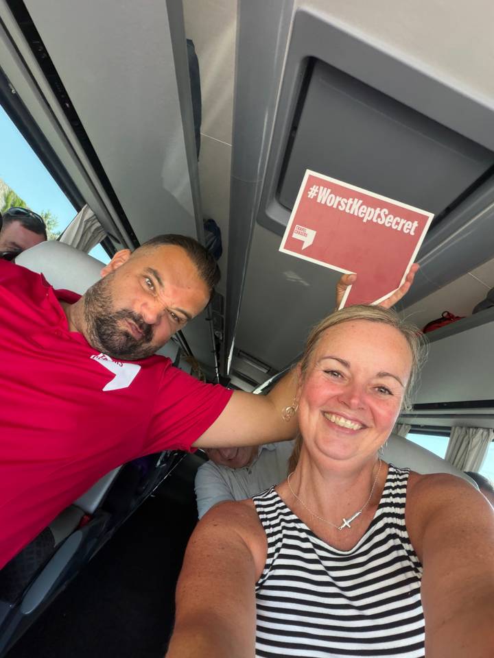 Two passengers joke around inside a tour bus, one holding up a playful sign while smiling for the camera.