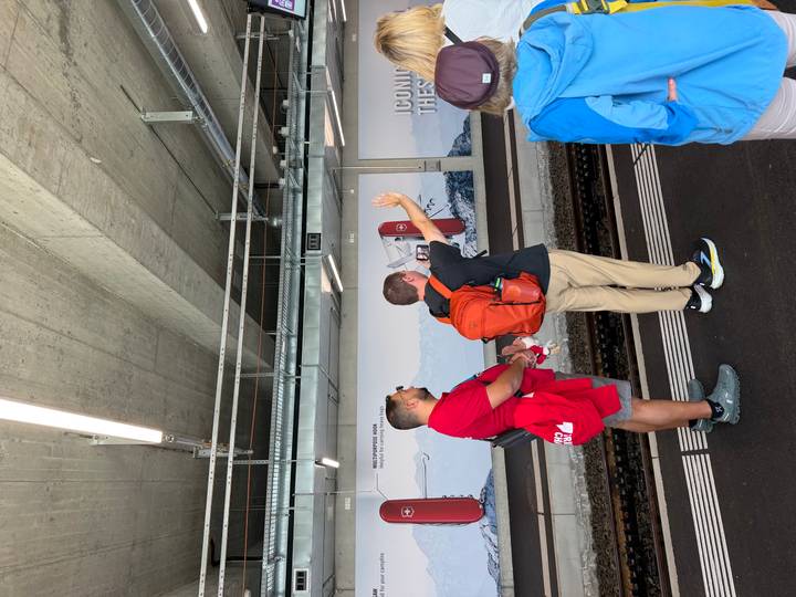 Travellers wait on a train platform beneath concrete ceilings and advertising panels featuring mountain imagery.