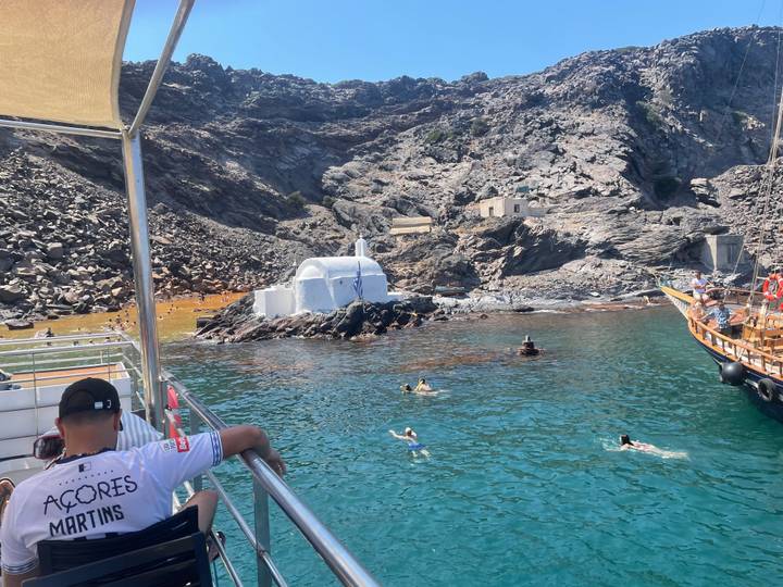 Swimmers enjoy turquoise water near a tiny white chapel on a rocky shore while visitors relax on a tour boat.