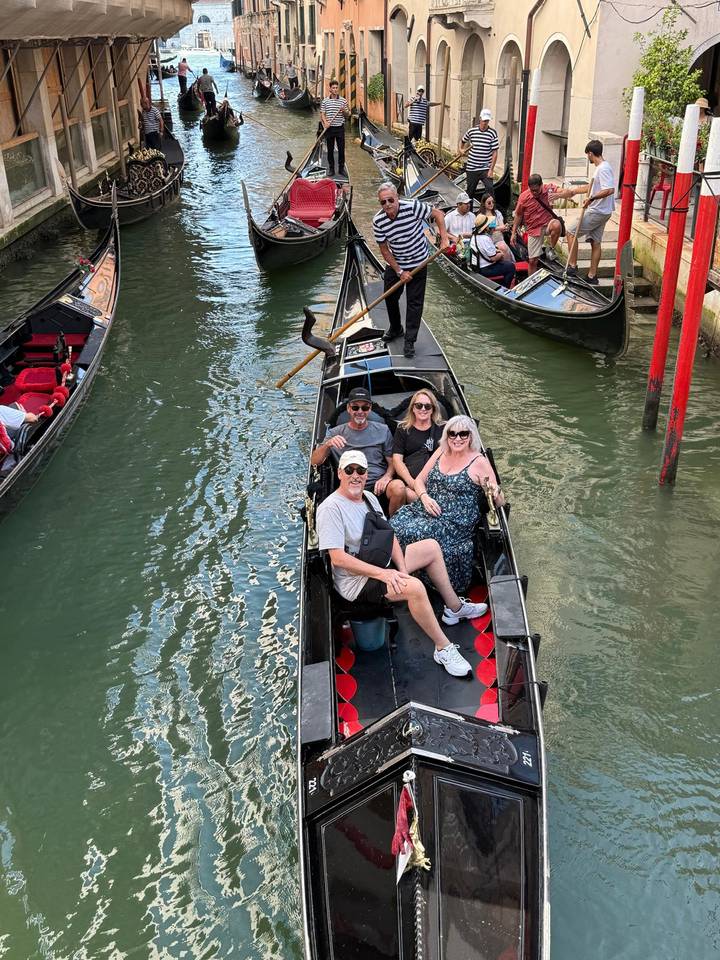 Four travelers sit smiling in a black gondola on a narrow Venetian canal.