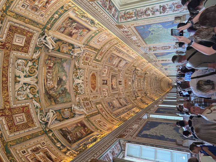 Crowded ornate gallery with a gilded vaulted ceiling and large maps lining the corridor walls.