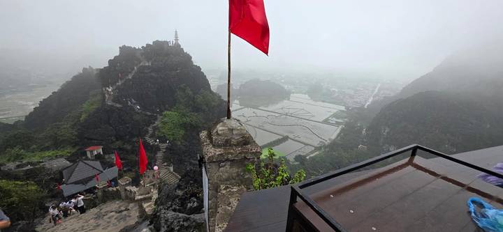 Foggy mountaintop at Hang Mua view with red flag and stepped path overlooking rice paddies.