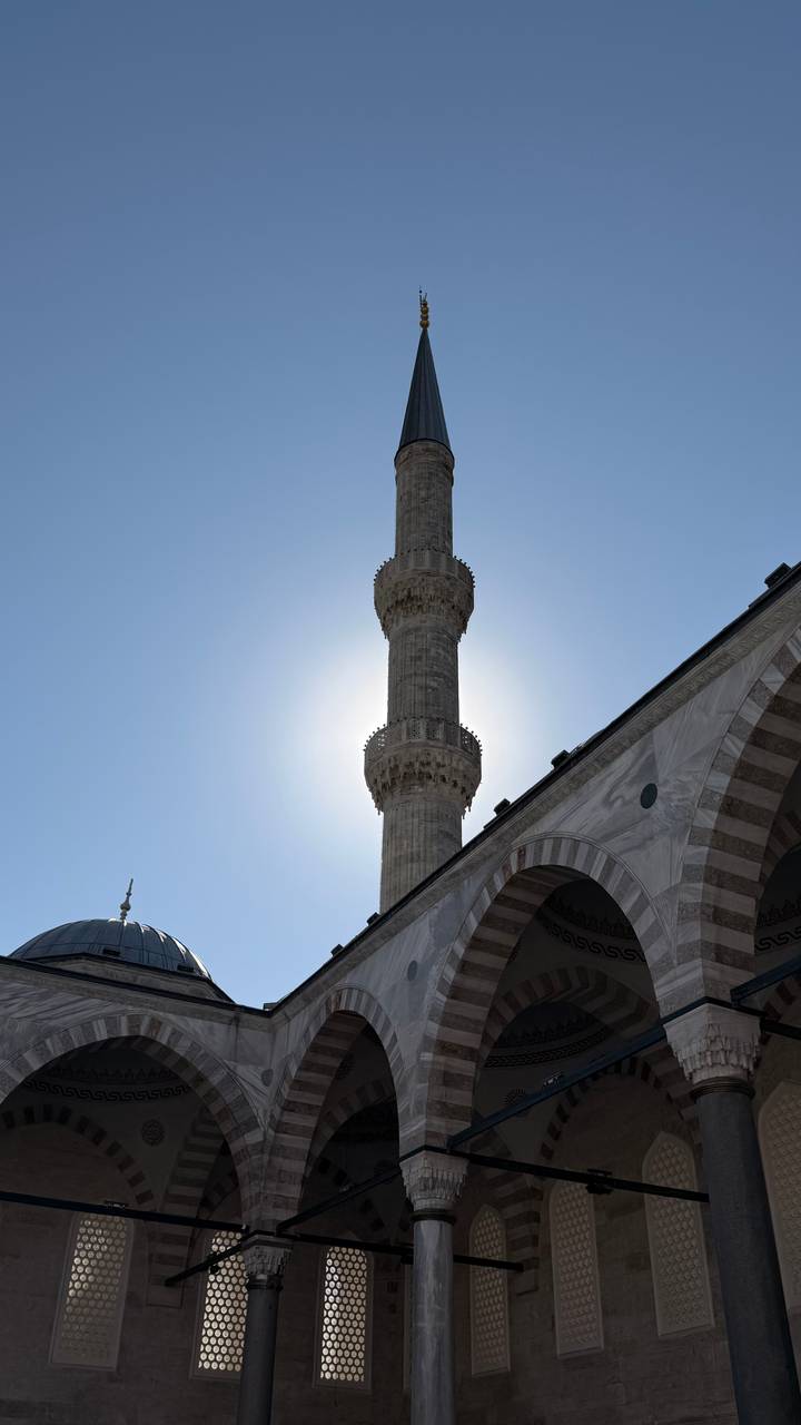 Tall stone minaret of a Turkish mosque silhouetted against the sun with clear blue sky.
