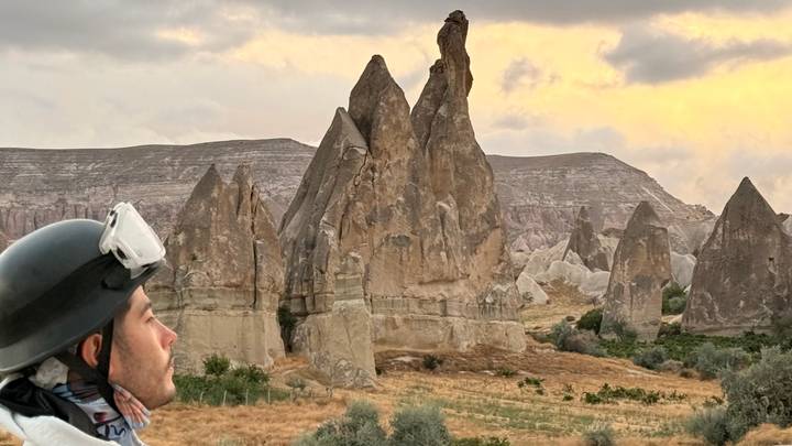 A traveler wearing a helmet gazes at dramatic fairy-chimney rock formations under a glowing evening sky in Cappadocia.