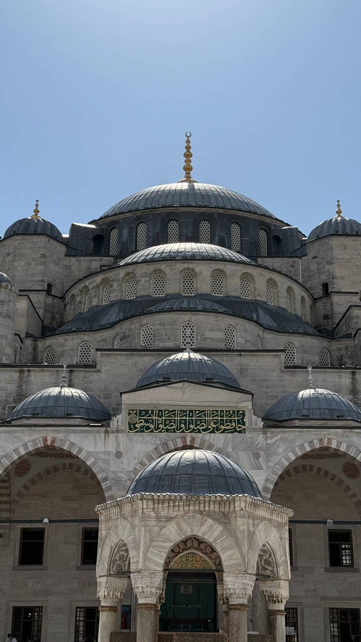 Close view of grey stone domes and stacked arches of a grand Ottoman mosque against a clear sky.