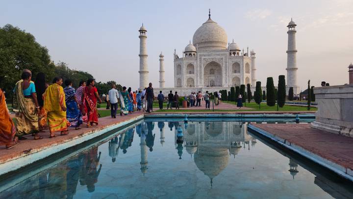 Visitors admire the Taj Mahal reflected perfectly in a blue pool during the warm glow of sunset.