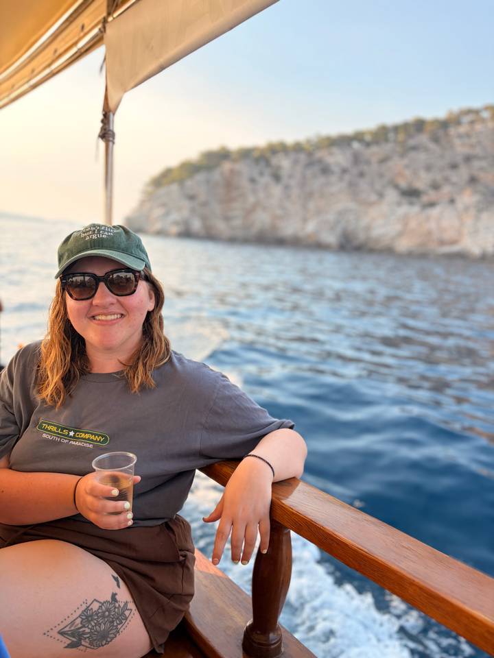 Smiling woman in sunglasses on a boat with deep-blue sea and rocky coastline behind her.