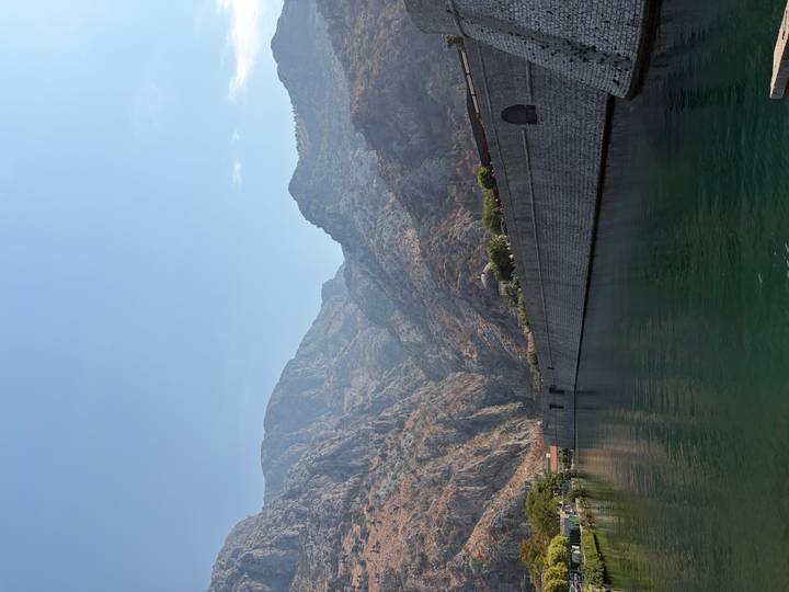 Steep rocky mountains rise above medieval stone ramparts reflected in calm green water.