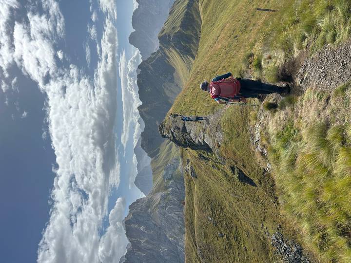 Hikers with backpacks trek along a narrow grassy ridge in high mountains with rugged peaks under a partly cloudy blue sky.