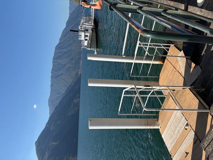 Pier with white mooring posts extending into a deep blue alpine lake with ferry boat and mountains