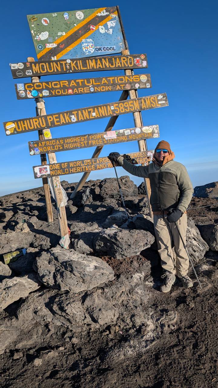 Climber in winter gear posing beside the Uhuru Peak sign atop Mount Kilimanjaro.
