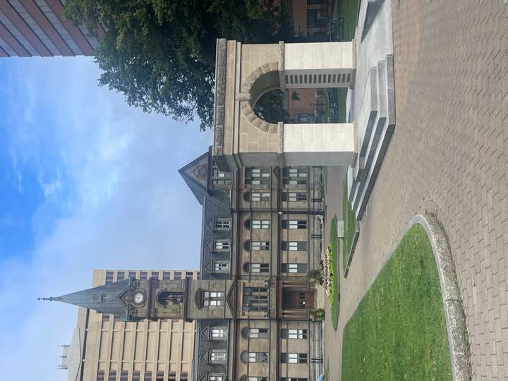 Historic sandstone civic building with clock tower beside a stone arch monument in a city square.