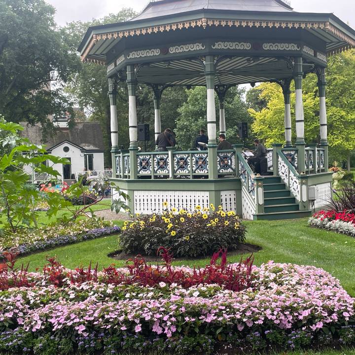 Musicians perform in a Victorian bandstand surrounded by colorful flowerbeds in a public garden.