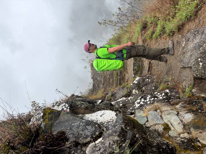 A lone hiker with a bright green backpack stands on a narrow stone trail carved into a mist-shrouded mountainside.