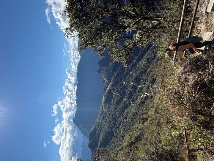 A woman descends metal steps overlooking a vast green valley and jagged Andean peaks under a clear blue sky.