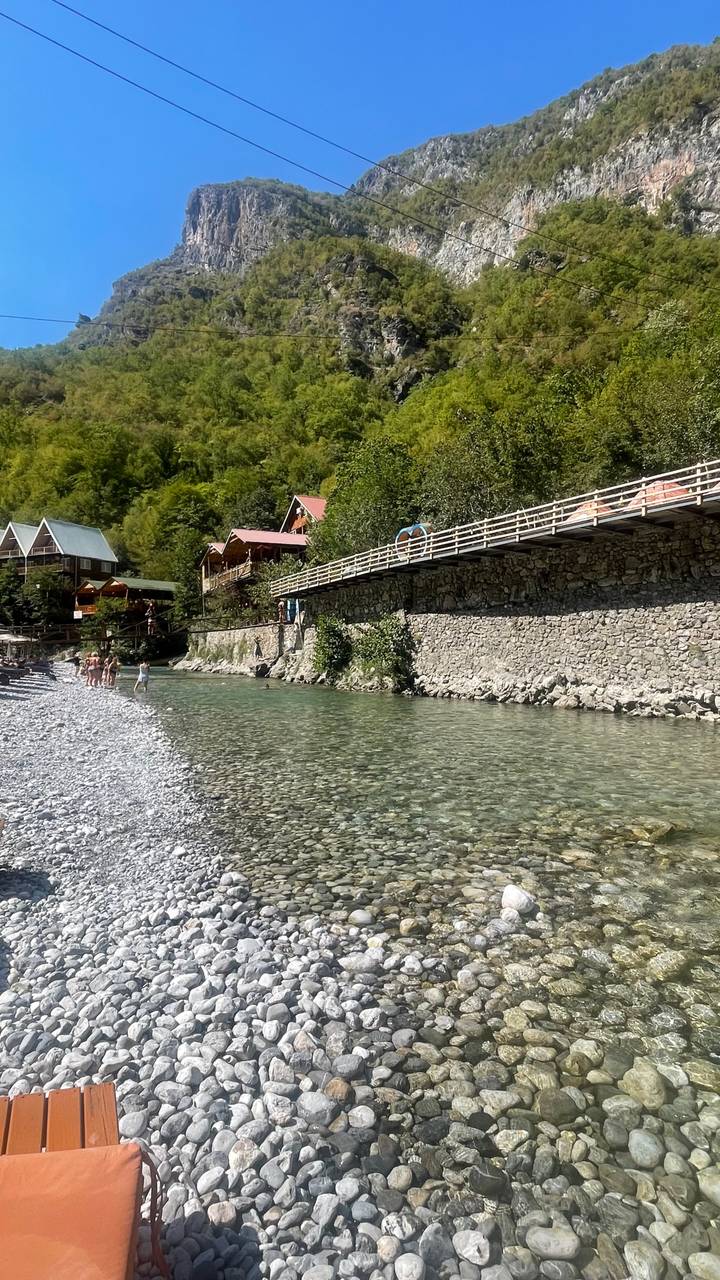 Clear turquoise river bordered by pebble beach and wooden cabins on a sunny day with bathers wading in the water.