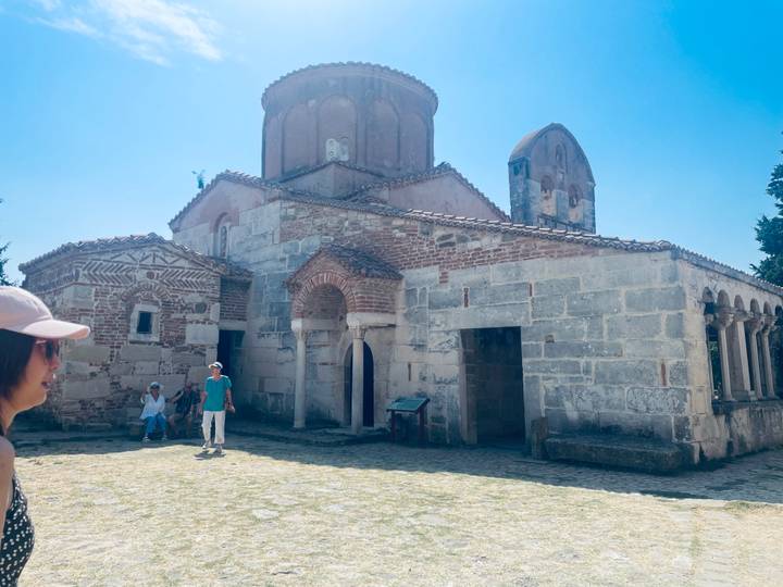 Tourists explore an old stone church with Byzantine architecture under a bright blue sky.