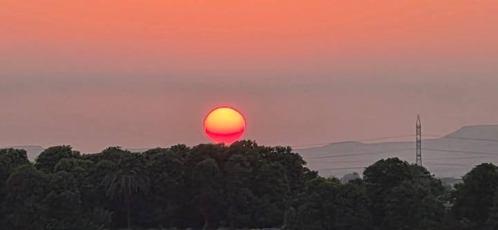Large orange sun setting behind silhouette of treeline and distant hills