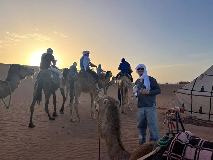 Travellers ride camels across desert dunes with the sun setting behind them near a Berber tent.