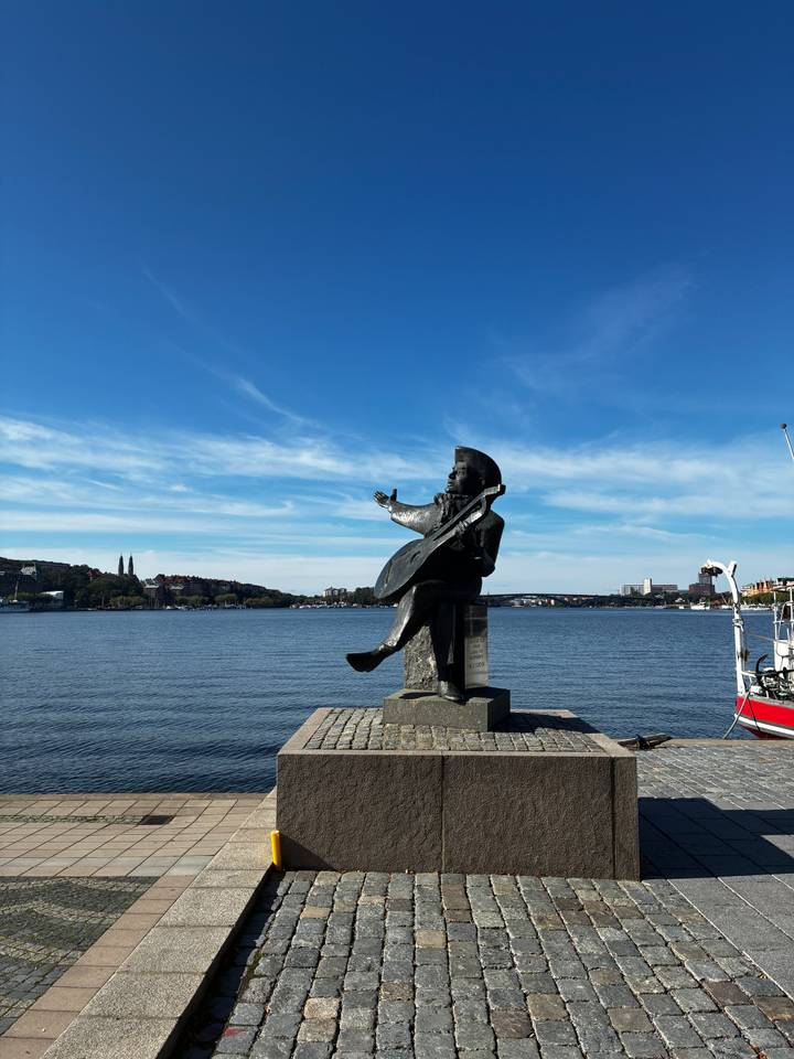 Waterfront bronze statue of a musician gestures toward Stockholm’s broad harbor under blue skies.