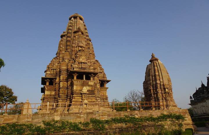 Sandstone spire and adjoining shrine of a Khajuraho temple rise against a clear blue sky.