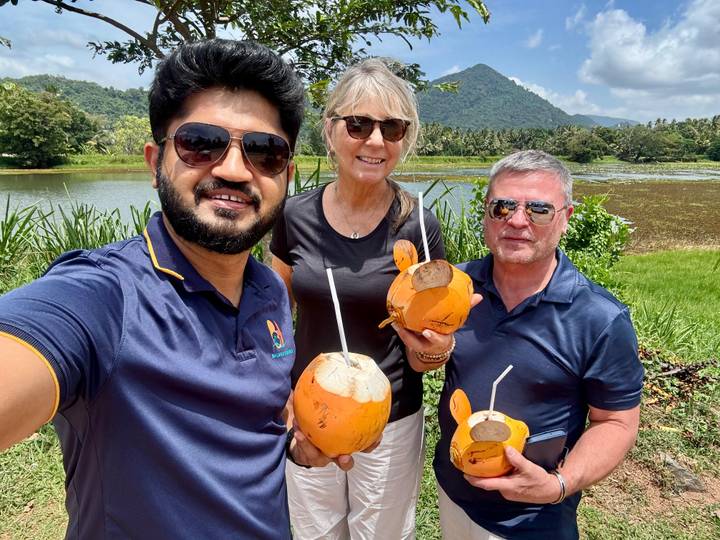 Selfie of guide and two travellers smiling while holding fresh coconuts beside a scenic lake with mountains.