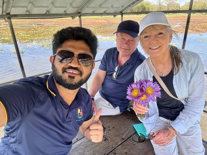 Selfie inside a small boat with guide and travellers holding purple lotus flowers over a lily-covered lagoon.