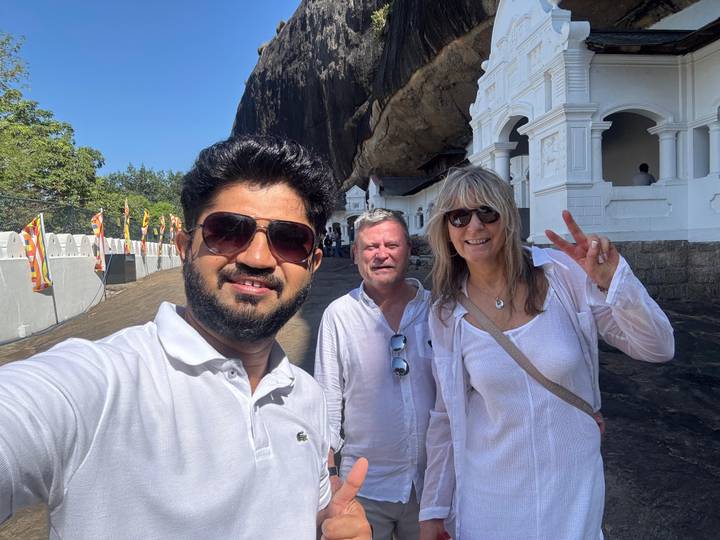 Selfie of guide and travellers outside a white-painted cave temple set beneath a towering rock cliff.