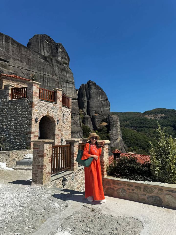 Traveler in a bright orange outfit posing beside a stone building with the towering rock pillars of Meteora in the background under a clear blue sky.