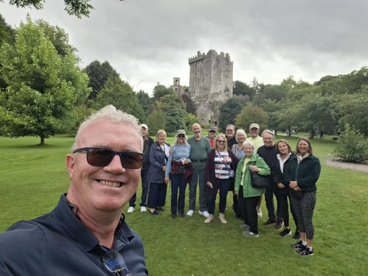 Tour group selfie on green lawn in front of stone Blarney Castle on cloudy day.
