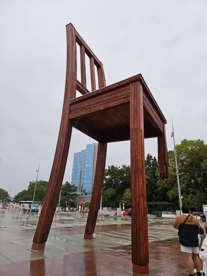 Giant wooden 'Broken Chair' sculpture stands tall near modern glass UN building against a grey sky