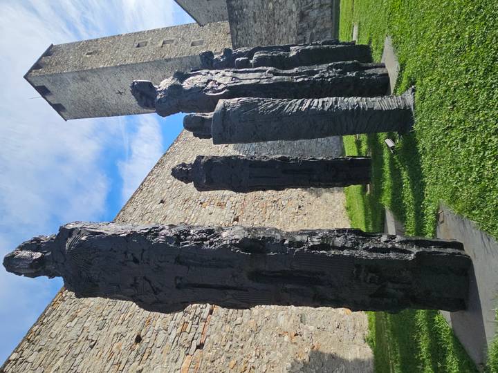 Row of tall, textured black sculptures stand beside an ancient stone wall and tower
