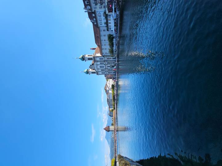 Lucerne’s Jesuit Church and Chapel Bridge tower reflect on calm Reuss River under a bright sky