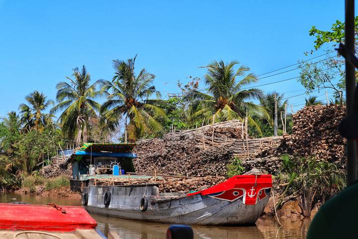 Small cargo boat moored along a Mekong Delta canal flanked by coconut palms and stacks of timber.
