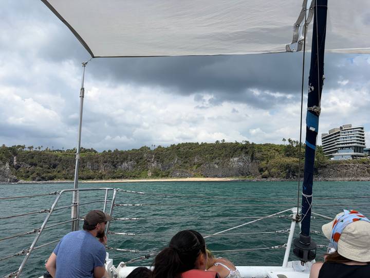 View from catamaran across emerald water toward forested volcanic coastline under patchy clouds.