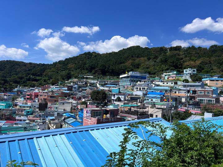 Colorful hillside village with dense pastel houses climbing a green slope beneath a bright blue sky.