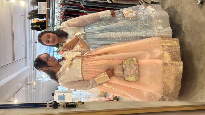 Two women wearing elegant hanbok dresses take a mirror selfie inside a rental shop.