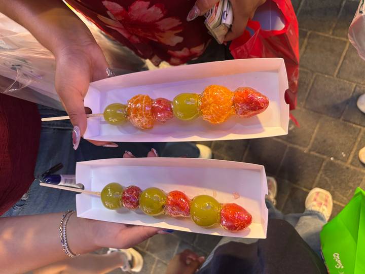 Two trays of colorful candied fruit skewers held up at a bustling night market.
