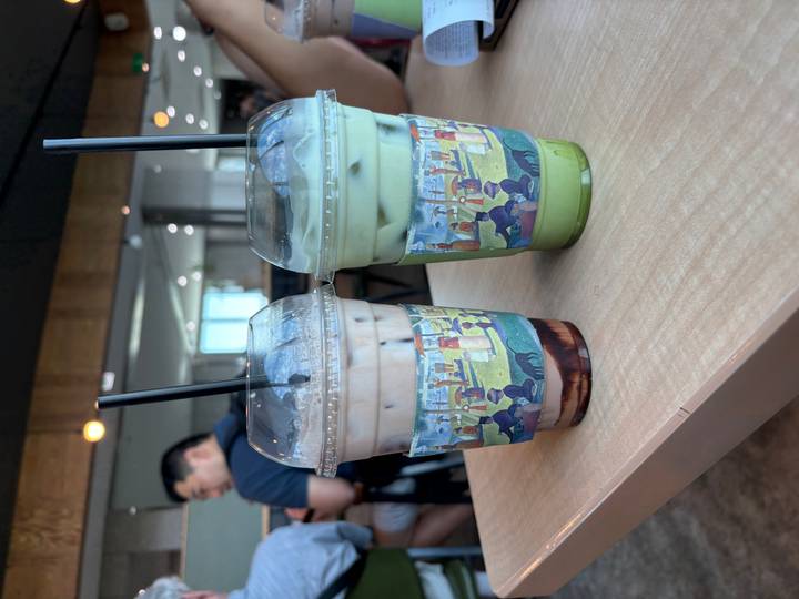 Two iced drinks, one chocolate and one matcha, rest on a cafe table with a patron blurred behind.