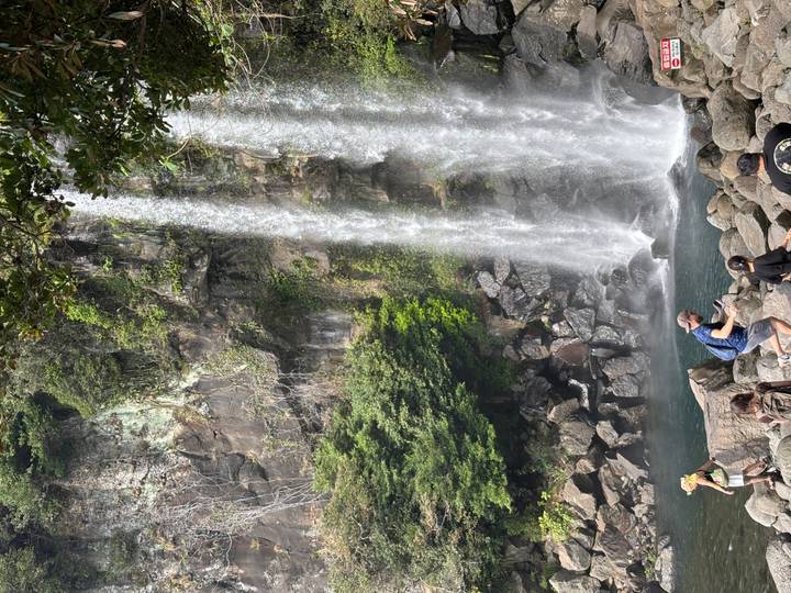 Visitors photograph a tall waterfall plunging into a rocky pool amid lush greenery.