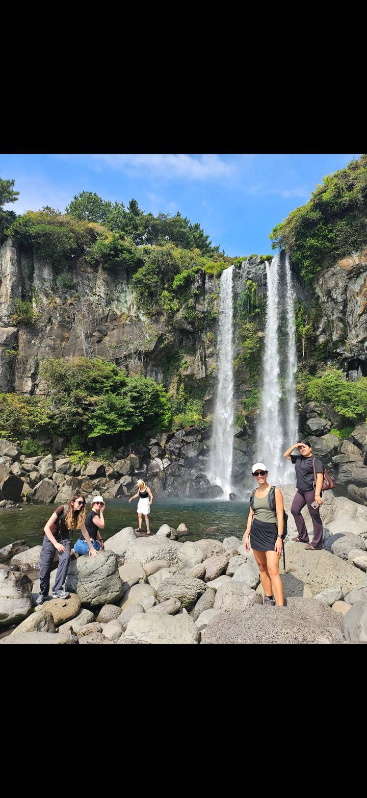 Twin waterfalls plunge over a rocky cliff into a misty pool where visitors pose on the stones below.
