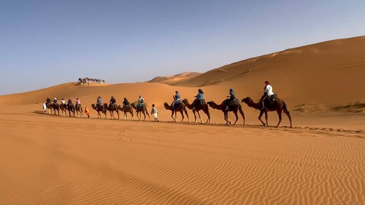 A long caravan of camel riders traverses rolling orange dunes under a clear sky.