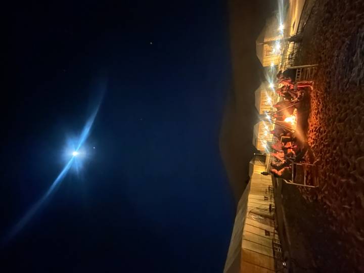 Nighttime desert camp where travellers sit around a glowing fire beneath a bright moon.